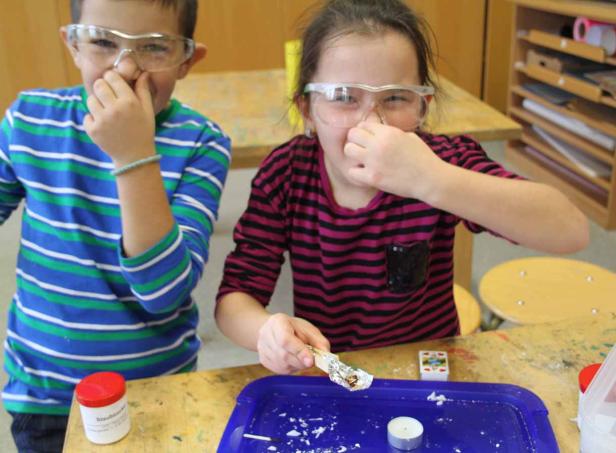 Zwei Kinder mit Schutzbrillen halten sich die Nase zu, während sie ein Experiment durchführen.