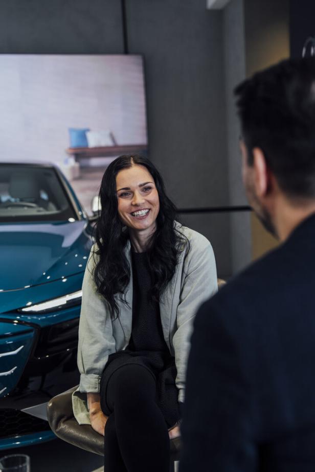 Eine Frau sitzt vor einem blauen Auto und lächelt einen Mann an.