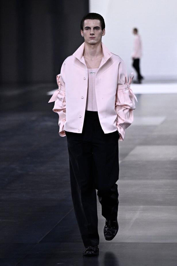 Ein Mann in einer rosa Jacke und schwarzen Hose läuft auf einem Laufsteg.