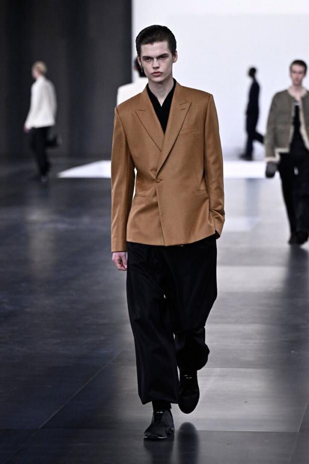Ein Mann in einem braunen Blazer und einer schwarzen Hose läuft auf einem Laufsteg.