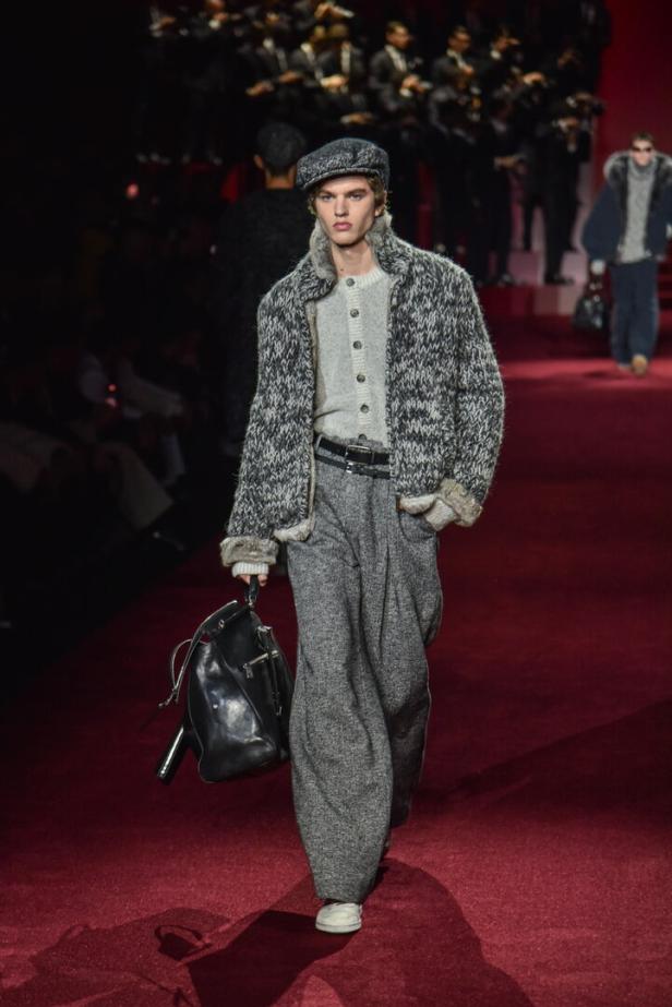Ein Mann in Tweedhose und Strickjacke mit einer schwarzen Ledertasche auf einem Laufsteg.
