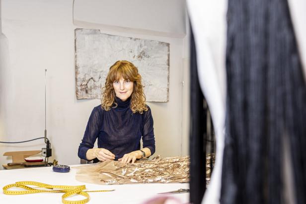 Eine Frau mit lockigem Haar sitzt an einem Tisch und näht an einem goldenen Stoff.