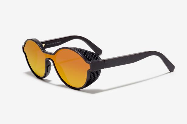 Eine 3D-gedruckte Sonnenbrille mit orangefarbenen Gläsern und Seitenschutz liegt auf einem weißen Hintergrund.