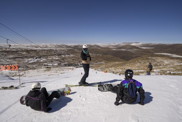 Zwei Snowboarder sitzen auf der Piste, ein Skifahrer steht. Blick auf eher wenig beschneite Berge.