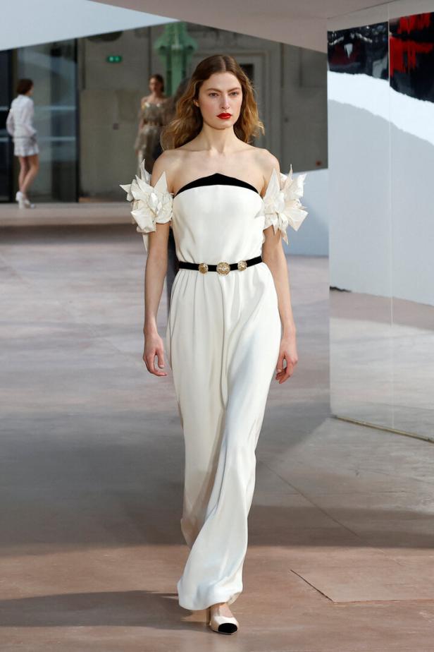 Eine Frau in einem weißen, bodenlangen Kleid mit floralen Details an den Schultern läuft auf einem Laufsteg.