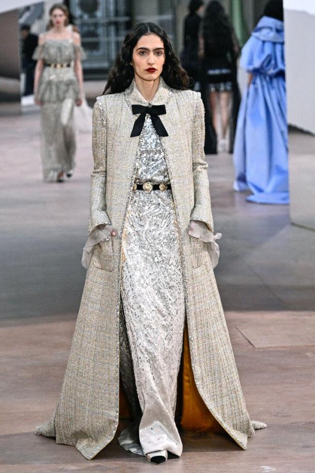 Eine Frau in einem langen, silbernen Paillettenkleid mit einem Tweed-Mantel auf einem Laufsteg.
