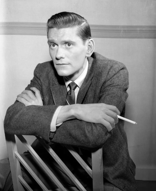 Ein Mann im Anzug lehnt an einem Stuhl und hält eine Zigarette.