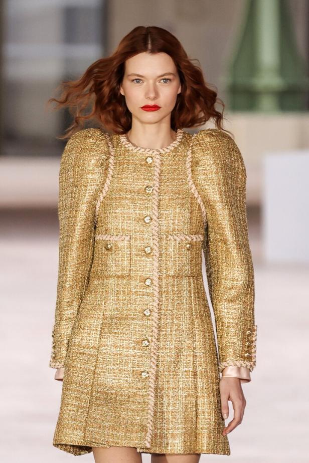 Ein rothaariges Model präsentiert ein goldenes Tweedkleid mit Puffärmeln und rosa Akzenten.