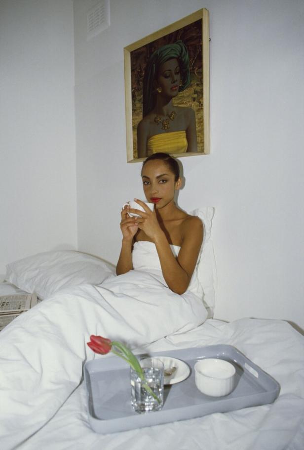 Sade sitzt im Bett und trinkt eine Tasse Tee; über ihr hängt ein Porträt.