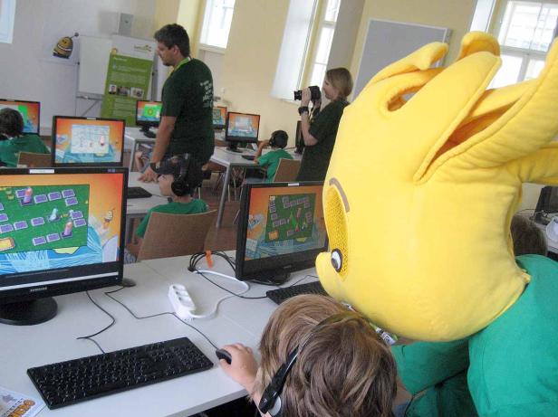 Kinder sitzen an Computern und spielen ein Lernspiel, während eine Person im gelben Kostüm zusieht.