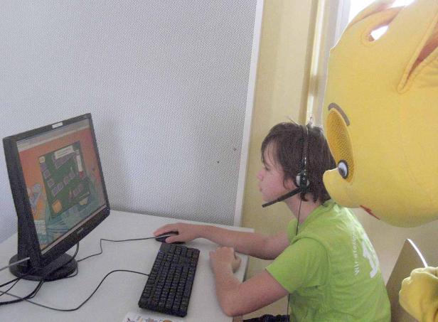 Ein Junge mit Headset spielt ein Computerspiel, während ein gelbes Maskottchen zusieht.