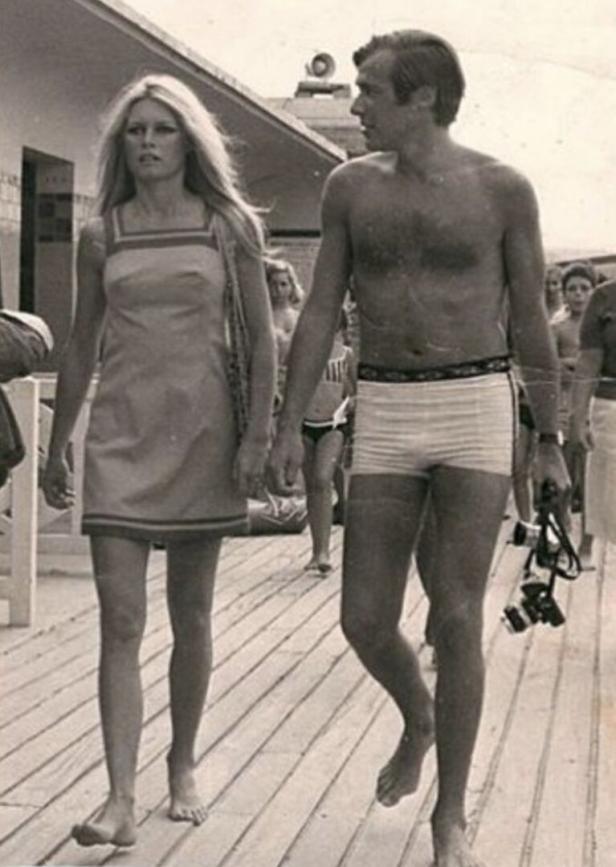 Brigitte Bardot und ein Mann mit Kamera gehen barfuß an einem Strand entlang.