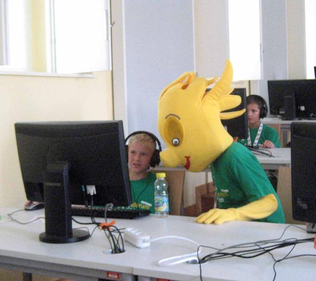 Ein gelbes Maskottchen und Kinder sitzen an Computern in einem Raum.