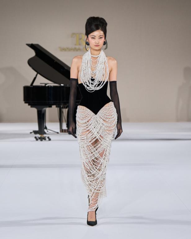 Eine Frau in einem schwarzen Kleid mit Perlen auf einem Laufsteg, im Hintergrund ein Klavier.