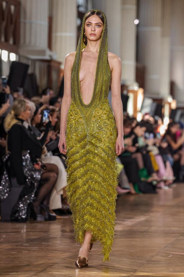 Ein Model präsentiert ein grünes, mit Perlen besetztes Kleid mit Kapuze auf dem Laufsteg.