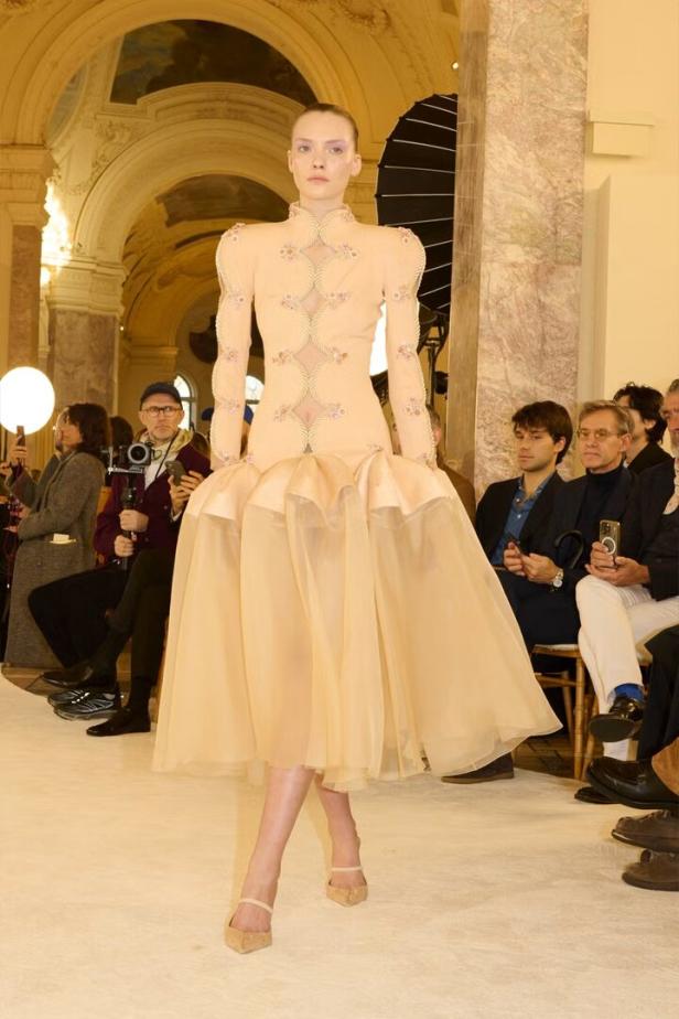 Ein Model präsentiert ein peachfarbenes Kleid mit langen Ärmeln auf einem Laufsteg.
