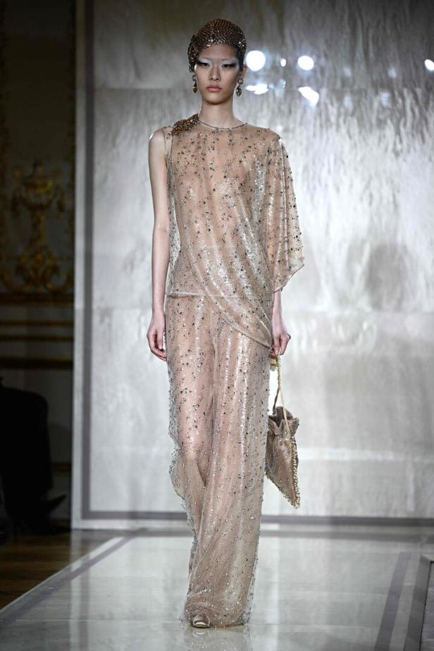 Ein Model präsentiert ein transparentes, paillettenbesetztes Ensemble mit passendem Kopfschmuck und einer Handtasche.
