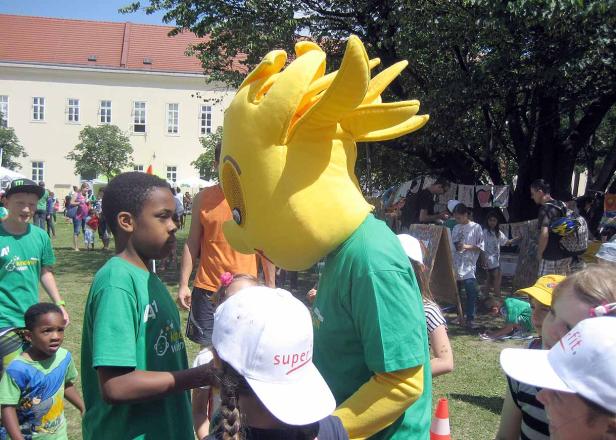 Ein gelbes Maskottchen interagiert mit Kindern auf einer grünen Wiese.