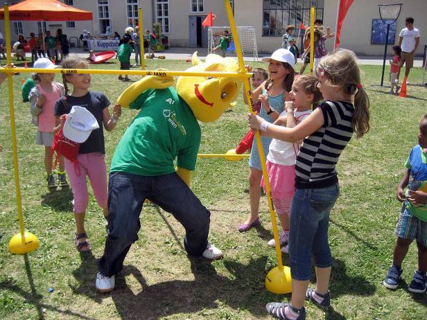 Kinder spielen Limbo mit einem gelben Maskottchen auf einer Wiese.