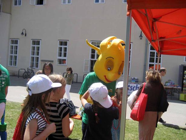 Eine gelbe Maskottchen-Figur interagiert mit einer Gruppe von Kindern im Freien.