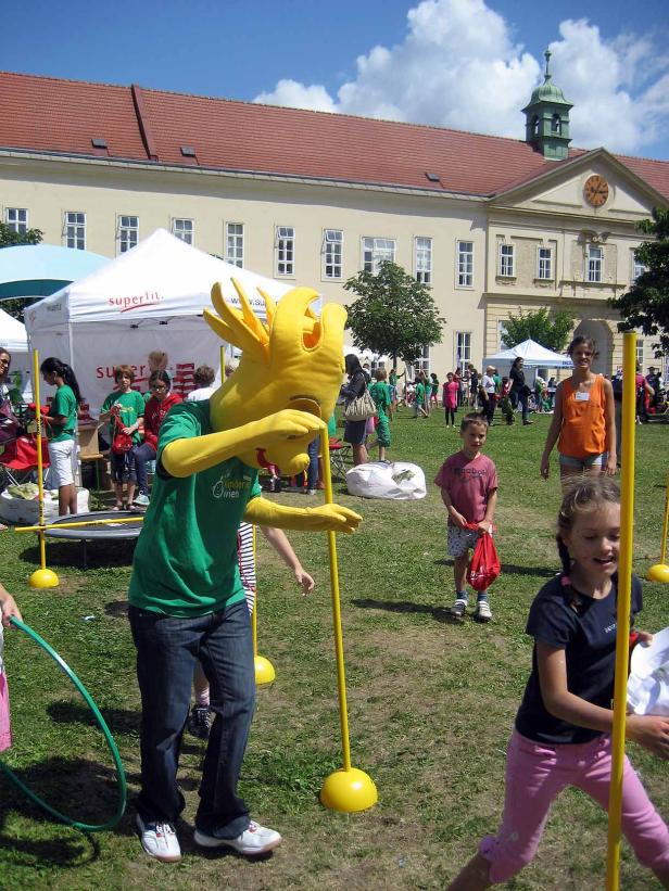 Ein gelbes Maskottchen und Kinder spielen auf einer Wiese mit gelben Stäben.