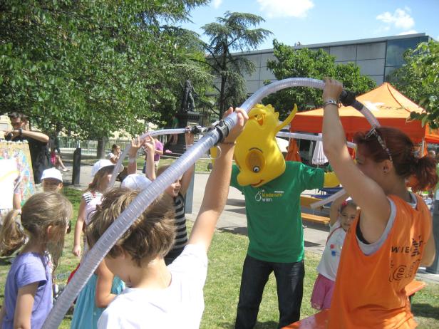 Kinder und ein Maskottchen halten einen Schlauch auf einer Veranstaltung von Kinderuni Wien.