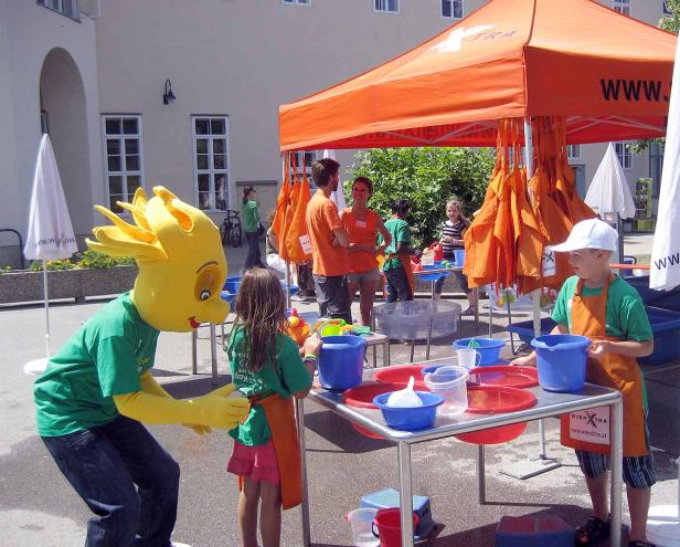 Ein gelbes Maskottchen und Kinder spielen mit Wasser und Eimern an einem Stand von wienXtra.