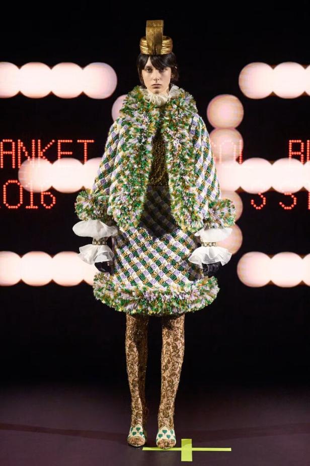 Ein Model präsentiert ein kariertes Tweed-Ensemble mit goldenen Leggings und Kopfschmuck auf einem Laufsteg.