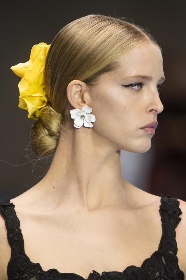 Eine Frau mit blonden Haaren trägt eine gelbe Blume und einen weißen Ohrring in Blütenform.