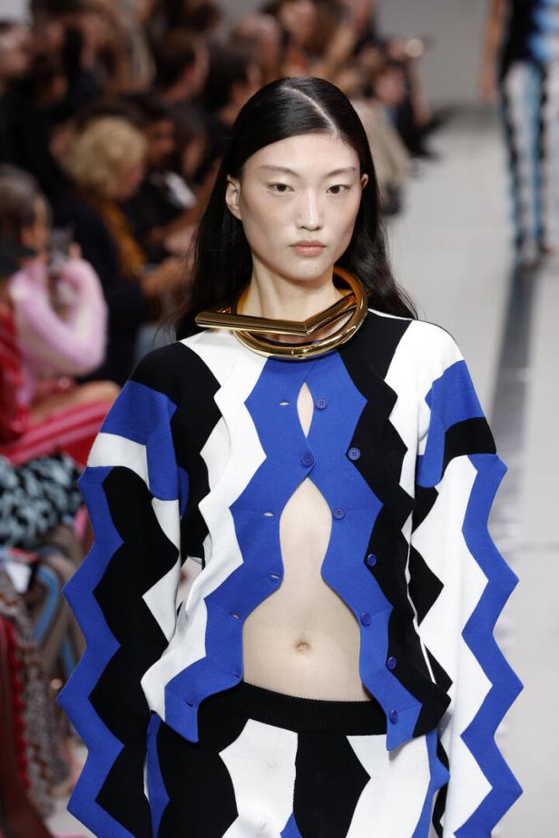Ein Model mit einem geometrischen Outfit und einem goldenen Halsreif auf dem Laufsteg.