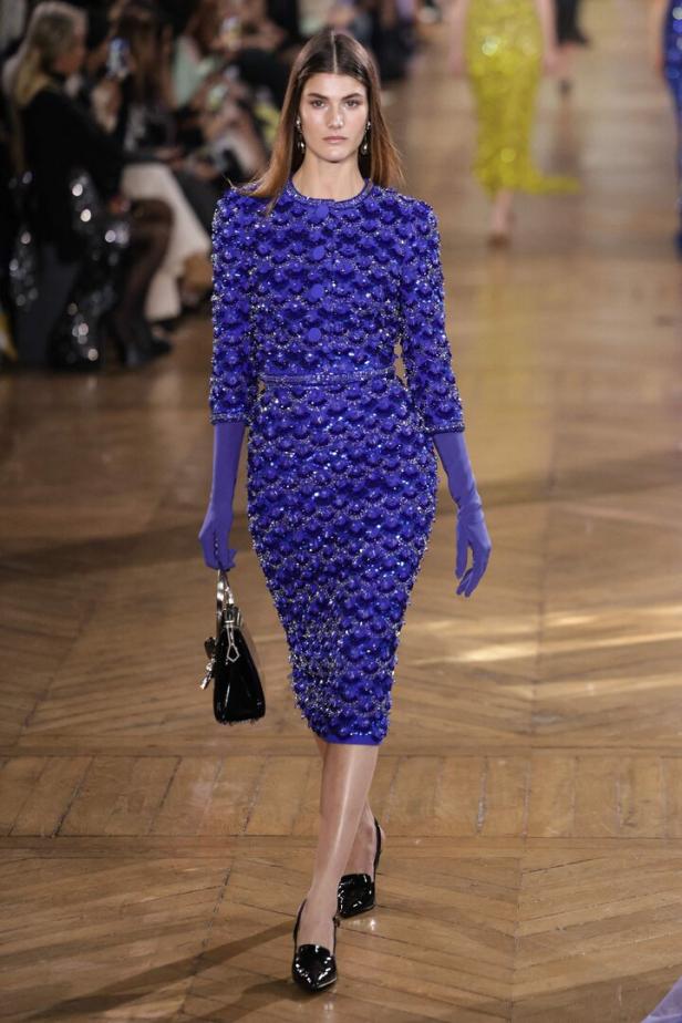 Ein Model präsentiert ein blaues, paillettenbesetztes Kleid mit passenden Handschuhen auf dem Laufsteg.