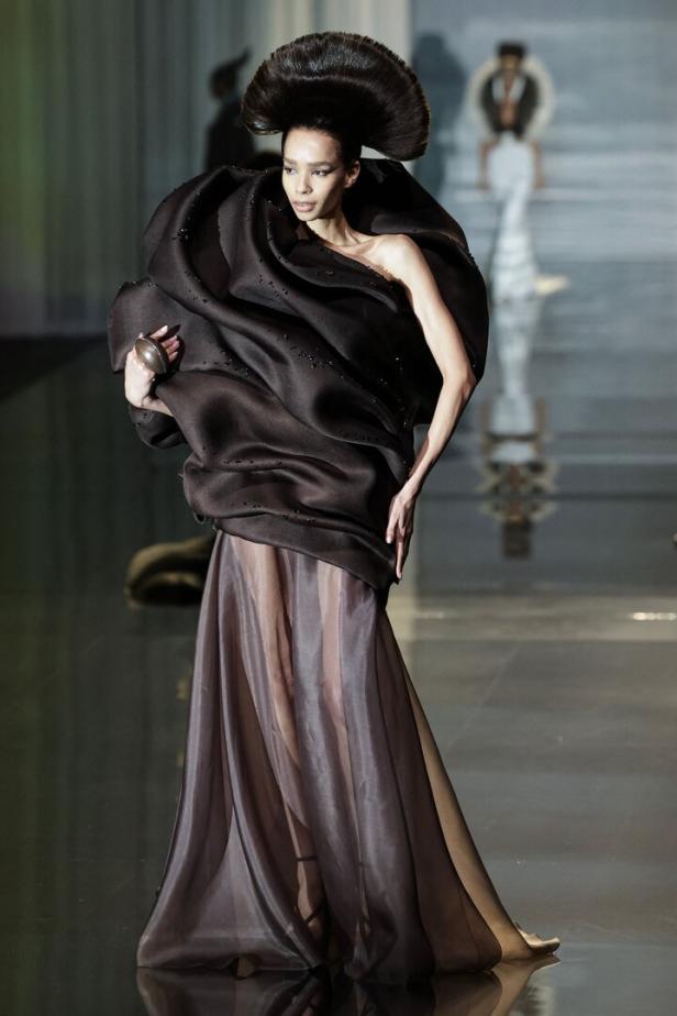 Ein Model präsentiert ein extravagantes, dunkles Kleid mit voluminöser Kragenform und auffälliger Frisur auf dem Laufsteg.