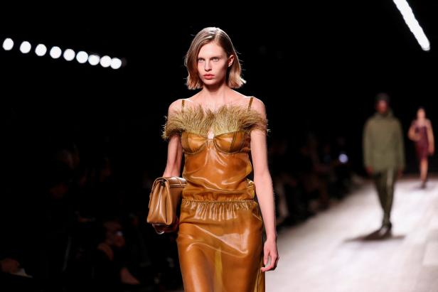 Ein Model präsentiert ein transparentes, ockerfarbenes Kleid mit Federn auf einem Laufsteg.