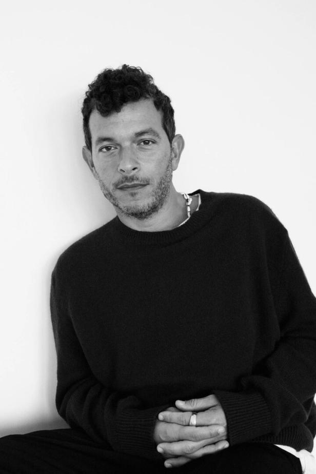 Ein Mann mit lockigem Haar und einem dunklen Pullover posiert vor einer weißen Wand.