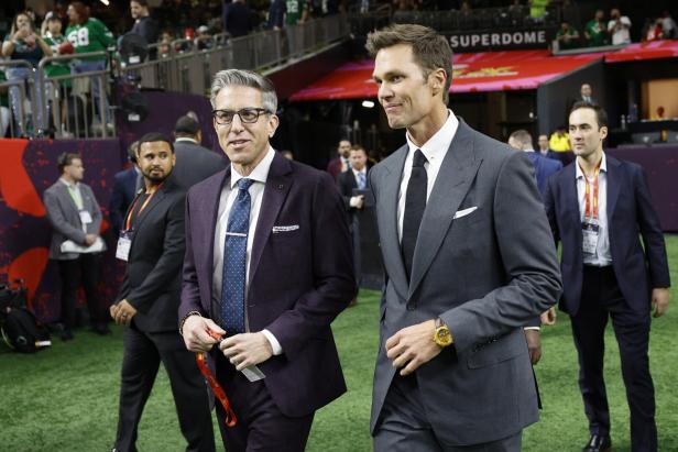 Tom Brady und ein weiterer Mann in Anzügen gehen über das Spielfeld.