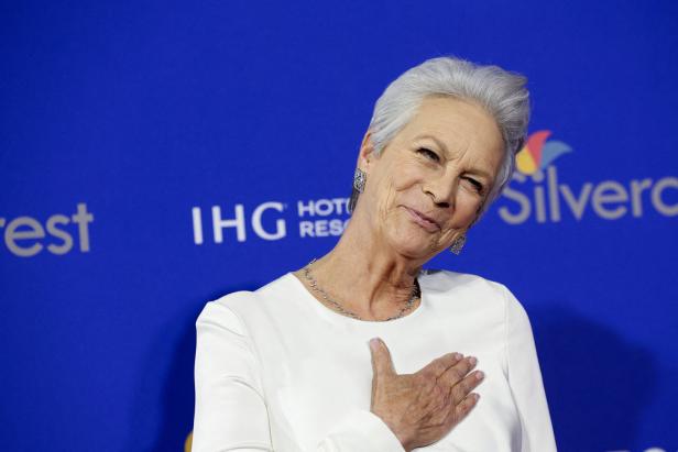 Jamie Lee Curtis lächelt und hält ihre Hand auf ihre Brust vor einem blauen Hintergrund.