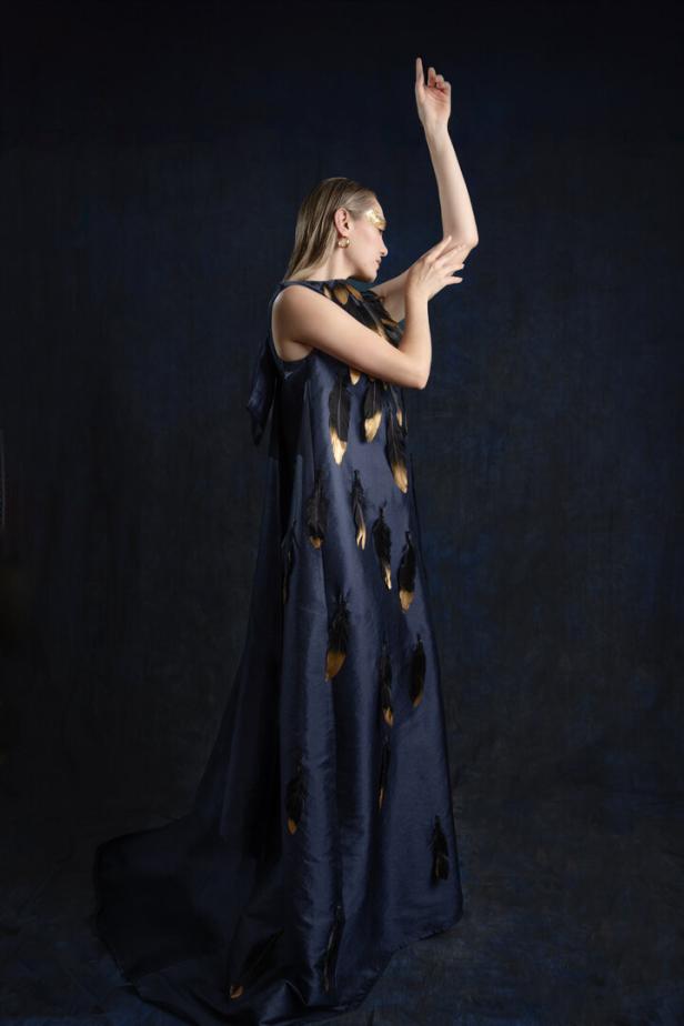 Eine Frau in einem langen, dunklen Kleid mit goldenen und schwarzen Federn posiert vor einem dunklen Hintergrund.
