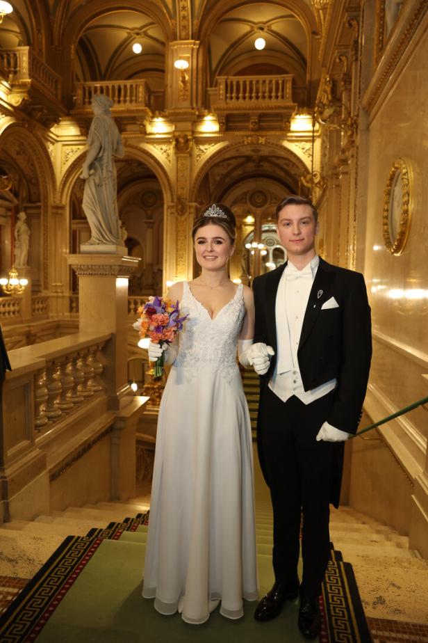 Ein Paar in formeller Kleidung steht auf einer Treppe in einem opulenten Gebäude.