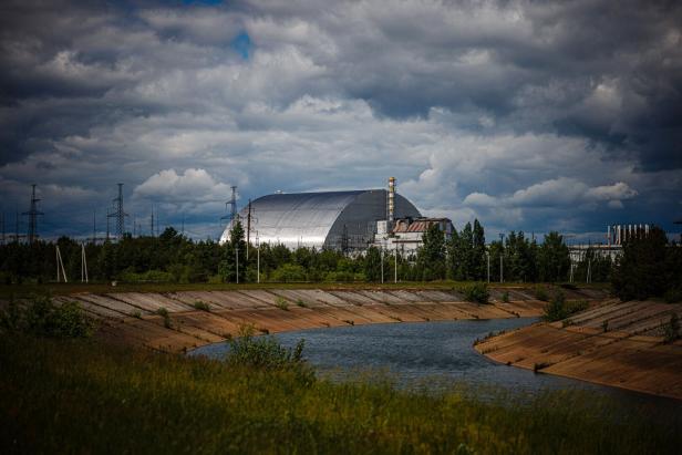 Der Sarkophag von Tschernobyl unter einem bewölkten Himmel, gesehen von einem grasbewachsenen Ufer aus.