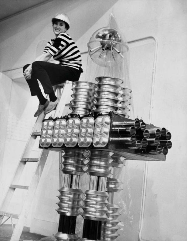 Eine Frau mit Schutzhelm sitzt auf einer Leiter neben einem Roboter mit einer Glaskugel als Kopf.