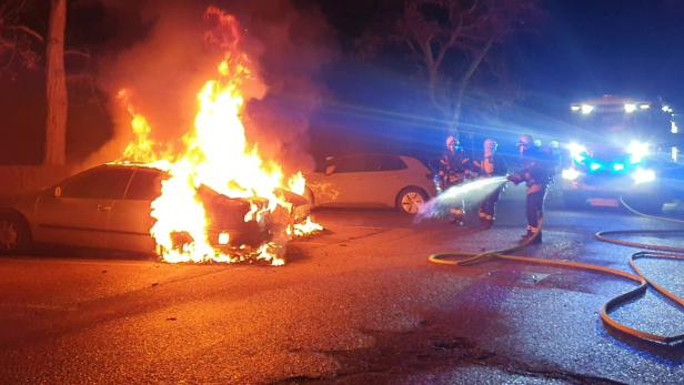 Feuerwehrmann mit Wasserschlauch und links ein Auto im Vollbrand im Hintergrund Blaulicht
