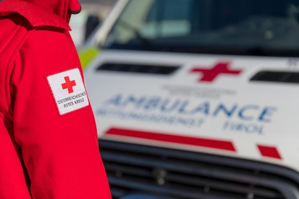 Eine Person in roter Jacke mit Rotkreuz-Emblem steht vor einem Rettungswagen mit rotem Kreuz.