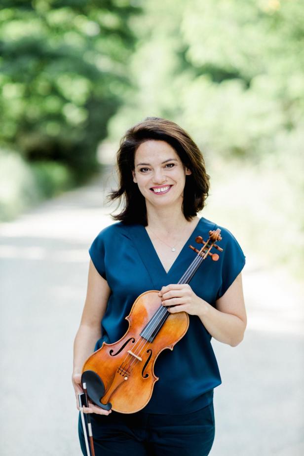 Eine brünette Frau steht da mit ihrer Violine in der Hand und lächelt.