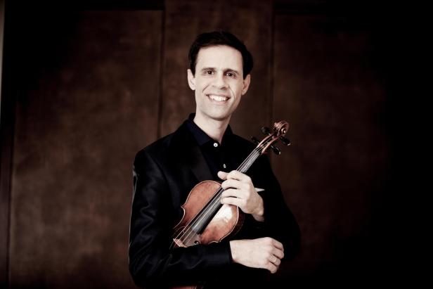 Ein Mann lächelt mit seiner Violine unter seinem Arm in die Kamera. 