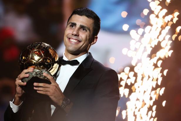 Ein lächelnder Mann im Anzug hält eine goldene Fußballtrophäe in den Händen.