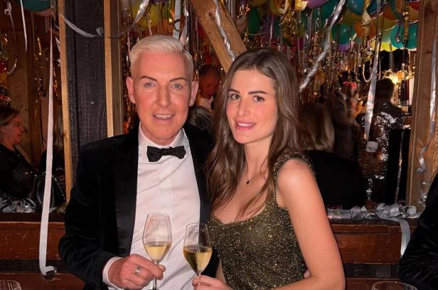 HP Baxxter: 60-jähriger blonder Mann sitzt mit einem Glas Sekt neben seiner 23-jährigen brünetten Freundin im Restaurant.