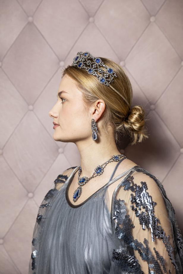 Eine Frau mit Tiara, Ohrringen und Halskette mit blauen Steinen vor einem rosa Hintergrund.