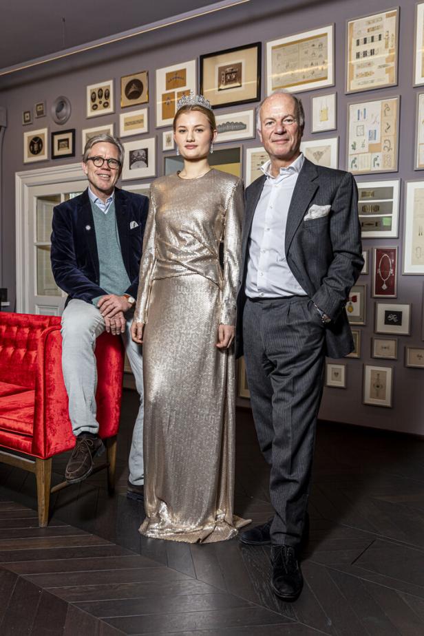 Eine junge Frau mit Tiara und goldenem Kleid posiert mit zwei Männern vor einer Wand mit Bildern.