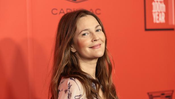 Drew Barrymore vor rotem Hintergrund und lächelt in die Kamera, der Kopf ist leicht schräg geneigt.
