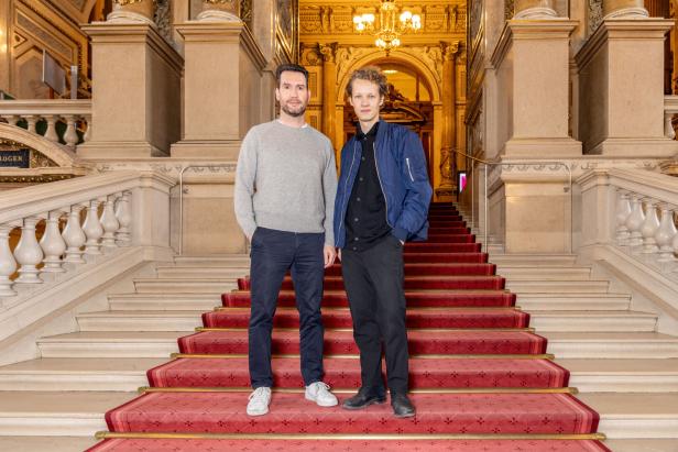 Zwei Männer stehen auf einer mit einem roten Teppich belegten Treppe in einem opulenten Gebäude.